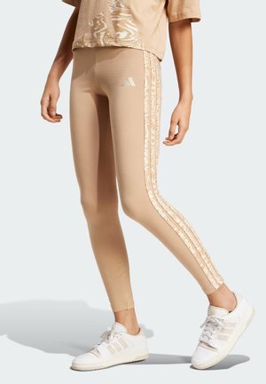 Leggings beige con un design aderente, caratterizzati da strisce laterali a fantasia e un logo adidas sull'anca. Indossati con sneaker bianche.