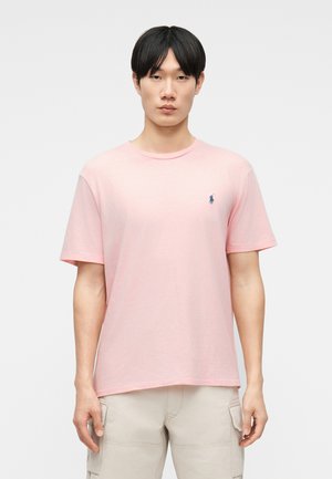 Young man wearing a light pink short-sleeve crew neck t-shirt with small blue logo on chest and beige cargo pants.