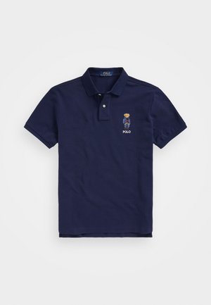 Granatowa koszulka polo z kołnierzykiem, dwiema guzikami i haftowanym misiem w dżinsach i czerwonym swetrze nad napisem "POLO" po lewej stronie klatki piersiowej.