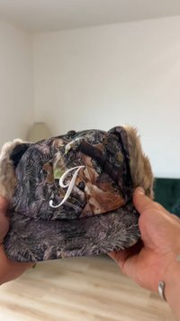 Camouflage cap featuring a fur trim and a white embroidered letter "J." The design includes varied earth tones and a textured surface.