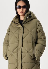 Olivgrüne Steppjacke mit Rautenmuster, Kapuze und frontaler Reißverschluss. Verfügt über große Taschen und Knopfakzente. Dunkler Pullover darunter.