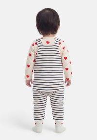 Παιδάκι που στέκεται με την πλάτη γυρισμένη, φοράει λευκή και ναυτικό ριγέ φορμάκι, κρεμ πουκάμισο με κόκκινα καρδούλες και ταιριαστά καλτσάκια σε λευκό φόντο.