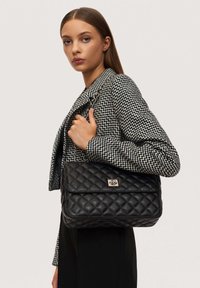 Quilted black leather handbag with a front flap and silver hardware accents, held by a person wearing a black-and-white patterned jacket.