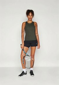 Mørkegrønn ermeløs topp kombinert med svarte treningsshorts, holde en tennisracket. Antrekket har et kroppsnært design og sporty estetikk.