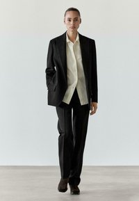 Czarna marynarka z wyraźnie zaznaczonymi ramionami, w połączeniu z jasnobeżową koszulą, dopasowanymi czarnymi spodniami i ciemnymi butami, na tle minimalistycznego wnętrza.