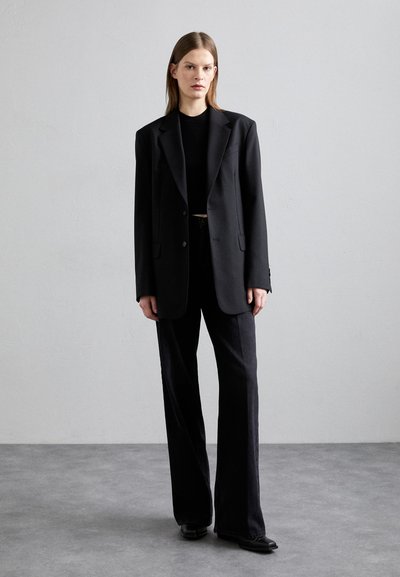 Blazer noir oversize porté sur un haut noir ajusté, associé à un pantalon large noir et des chaussures noires élégantes. Tissu lisse et structuré.