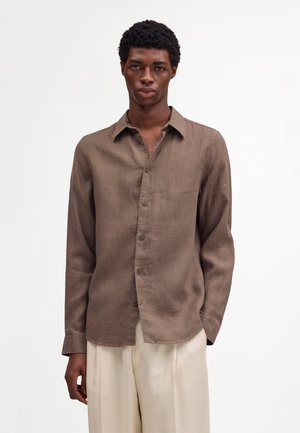 Młody mężczyzna ubrany w brązową koszulę na guziki z długim rękawem oraz luźne, jasnobeżowe spodnie, stojący na tle jednolitego białego tła.