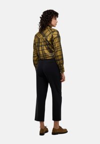 Chemise à carreaux jaune et noire, design court, à boutons, associée à un pantalon noir ajusté et des chaussures brunes. Fond uni.