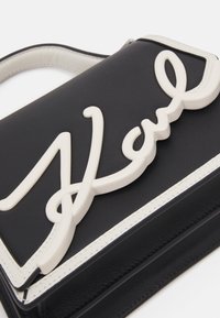 Czarna skórzana torba z białym elementem podpisowym. Charakteryzuje się schludnym, uporządkowanym designem oraz kontrastowym przeszyciem wzdłuż brzegów.