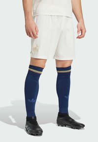 Witte sportieve shorts met een subtiel logo, gecombineerd met blauwe kniekousen met meerkleurige strepen en zwarte voetbalschoenen.