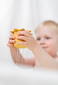 Geel plastic gietspeeltje dat met beide handen wordt vastgehouden, met een geschulpte rand; water stroomt vanaf de onderkant naar beneden, tegen een onscherpe achtergrond.