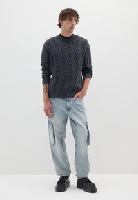 Dunkelgrauer Kabelstrickpullover, hellblaue Cargo-Jeans, schwarze Plateauschuhe. Das Modell steht vor einem weißen Hintergrund und zeigt eine lockere Passform.