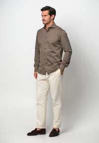 Camicia marrone a maniche lunghe con bottone frontale, abbinata a pantaloni bianchi e mocassini in camoscio scuro, creando un contrasto di colore e texture.