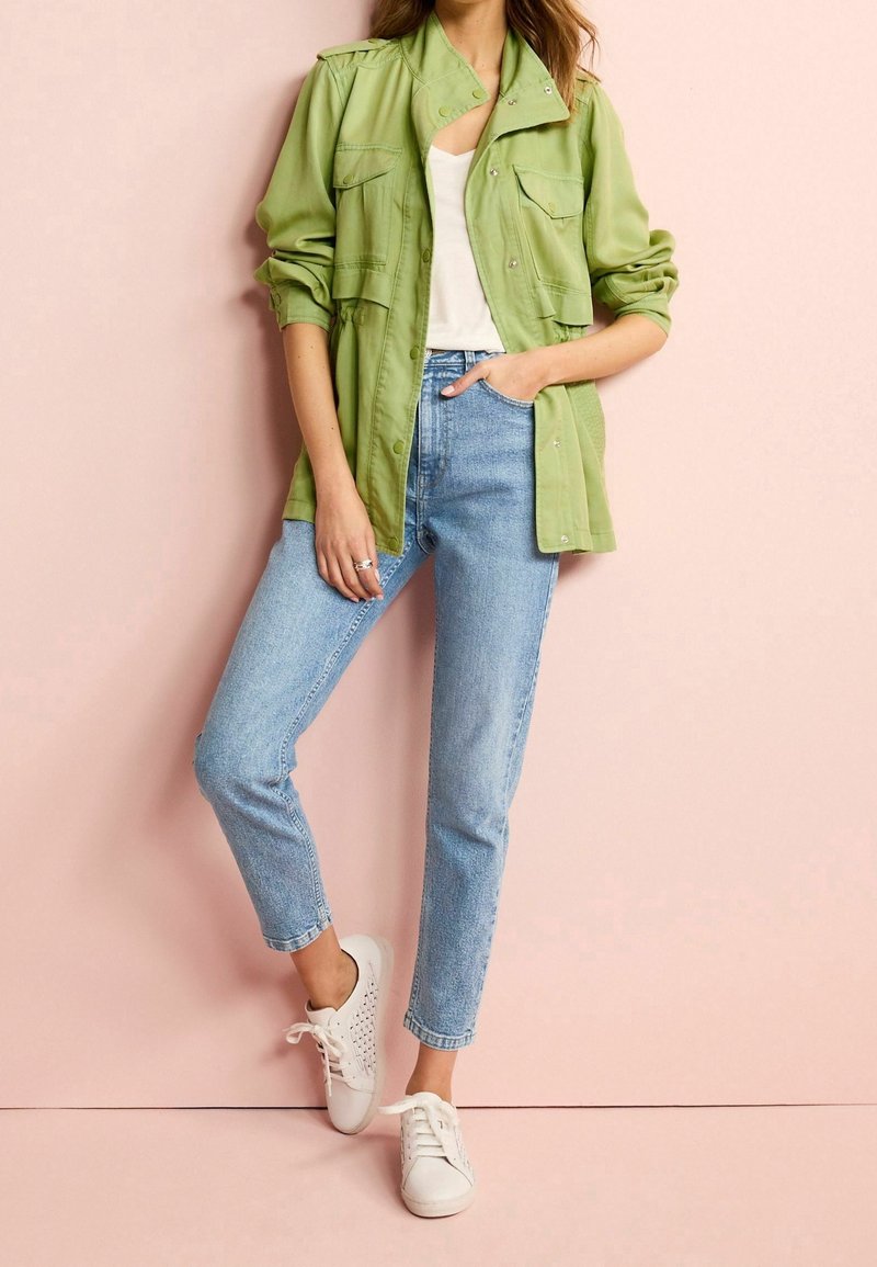 Femme portant une veste vert clair, un haut blanc, un jean bleu clair et des baskets blanches, posant devant un mur rose pâle.