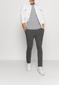 Homme portant une veste en denim blanche, un t-shirt rayé noir et blanc, un pantalon gris foncé, des baskets blanches, se tenant les mains dans les poches.