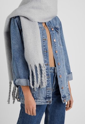 Personne portant une écharpe à franges gris clair sur une veste en denim bleu et un jean, avec un haut beige en dessous.