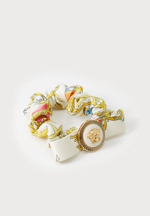 Scrunchie hecho de tela blanca con un estampado floral colorido. Presenta un charm redondo dorado con un diseño de cabeza de león en el centro.