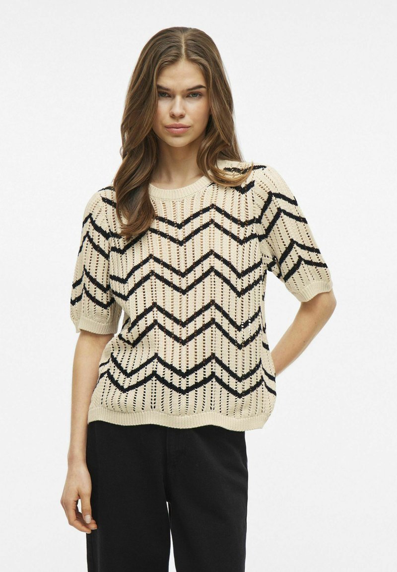 Strickpullover in Creme mit schwarzem Zickzackmuster. Verfügt über kurze Ärmel, einen runden Halsausschnitt und eine lockere Passform. Texturiertes, leichtes Material.