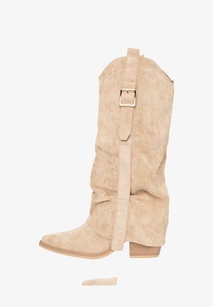 Beige suède enkellaarsjes met een nonchalant ontwerp, puntige neus, blokhak en een riem met metalen gespsluiting aan de buitenkant.