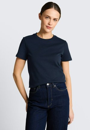 Frau mit braunem, zurückgebundenem Haar, die ein einfarbiges, marineblaues T-Shirt mit kurzen Ärmeln und eine dunkelblaue, hoch taillierte Jeans vor einem grauen Hintergrund trägt.