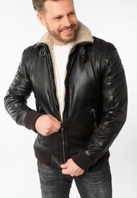 Chaqueta bomber de cuero negro con un collar de piel de oveja en color marrón claro. Presenta un cierre con cremallera, bolsillos frontales y puños con tejido acanalado. Textura suave.
