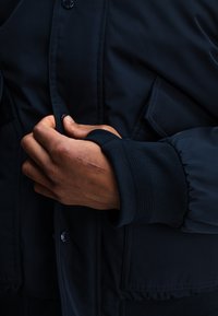 Marineblauer Bomberjacke mit gerippten Bündchen, einer Knopfleiste und seitlichen Taschen. Glatte Stoff mit dezentem Glanz und minimalen Steppdetails.