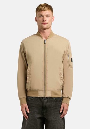 Ung mann iført en beige bomberjakke med glidelås, ribbestrikkede mansjetter og nederkant, står foran en ensfarget hvit bakgrunn.