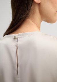 Licht beige blouse met een gladde textuur, voorzien van een sleutelgatopening aan de achterkant, bevestigd met een ronde metalen knop.