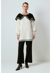 Abito in maglia color crema con colletto nero a ondina, maniche lunghe e rifiniture nere, abbinato a pantaloni neri larghi e stivaletti a punta.