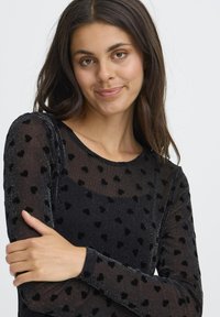 Sort mesh-top med lange ærmer, prydet med fløjelshjerteformede detaljer og en subtil glans. Rundet halsudskæring og tætsiddende silhuet.