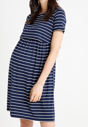 Femme enceinte portant une robe de maternité manches courtes rayée horizontale bleu marine et blanche, debout sur un fond blanc.