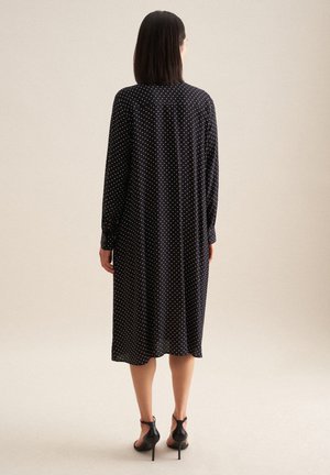 Abito nero con pois, maniche lunghe e scollatura alta, che scende sotto il ginocchio, abbinato a scarpe con tacco alto nere.