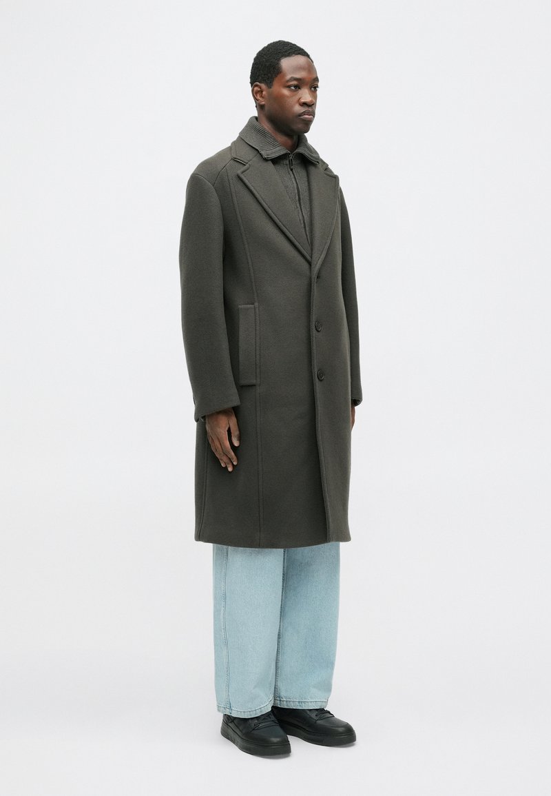 Olijfgroene jas met een gestructureerd ontwerp, enkelvoudige knopen en een gestructureerde kraag, gecombineerd met lichtblauwe wijde jeans en zwarte schoenen.