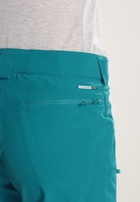 Tealfärgade vandringsbyxor tillverkade av lättviktsmaterial. Har en ficka med dragkedja bak, sydda detaljer och en figurnära midja med elastiskt snöre.