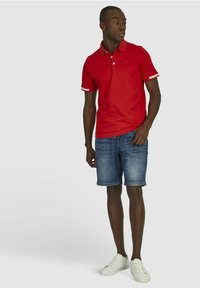 Rode polo-shirt met een kraag en drie knopen, gecombineerd met denim shorts en witte sneakers. De polo heeft contrasterende gestreepte accenten op de mouwen.