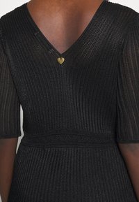 Personne à la peau foncée portant une robe en maille texturée noire avec un décolleté en V dans le dos et une petite étiquette dorée en forme de cœur près du cou.