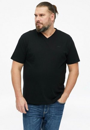 Bearded man with hair tied back wears black short-sleeve V-neck shirt and blue jeans, standing with one hand in pocket, looking left.