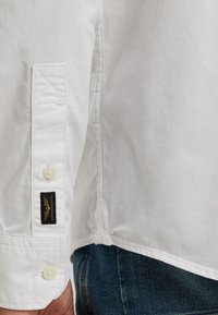 Camisa blanca de botonadura con puños reforzados, etiqueta, costura sutil y dobladillo recto, combinada con jeans de mezclilla azules.