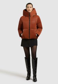 Giubbotto imbottito color ruggine con un ampio cappuccio, chiusura a zip e due tasche frontali. Le maniche sono leggermente a sbuffo; indossato sopra una minigonna nera.