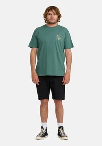 Mintgrøn bomulds t-shirt med korte ærmer og et lille flerfarvet blomsterprint på venstre bryst, parret med sorte shorts og sorte sneakers.