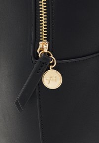 Detail černé kožené tašky s pozlaceným zipem a pozlaceným přívěskem s logem značky. Hladký povrch s jemným prošíváním a minimalistickým designem.