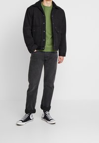 Černá denimová bunda s beránkovým límcem, zelená mikina, tmavě šedé d�žíny a černé tenisky Converse. Představuje uvolněný vrstvený vzhled.