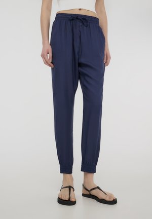 Granatowe, lekkie spodnie dresowe z elastycznym pasem i sznurkiem, zakończone ściągaczami, o gładkiej fakturze. Uzupełnione czarnymi klapkami.