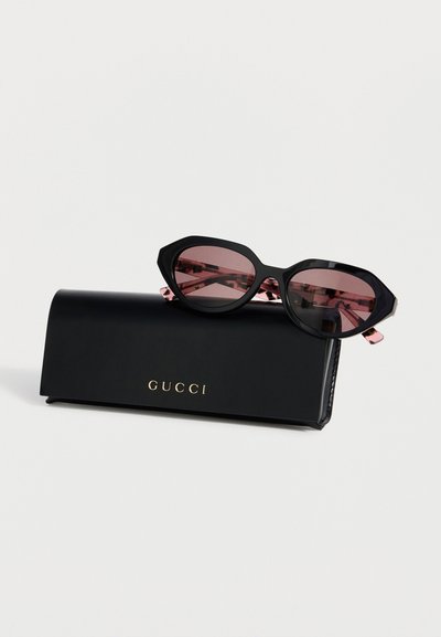 Lunettes de soleil œil de chat noires avec verres roses reposant sur un étui Gucci noir avec logo doré sur un fond blanc uni.