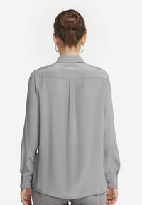 Hellgraue Bluse mit langen Ärmeln, glatter Textur, geknöpften Manschetten und gerundetem Saum. Verfügt über eine Naht im Rückenbereich und dezente Faltendetails.