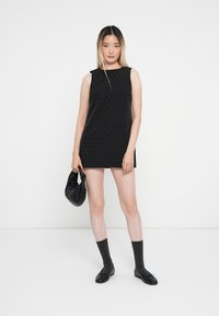 Rochie neagră fără mâneci, cu un model de puncte argintii, tiv drept, asociată cu pantofi negri și șosete gri până la gleznă, purtată cu o geantă neagră.