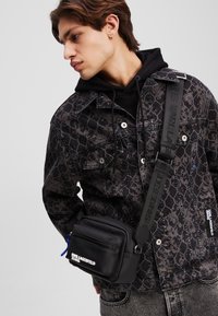 Sort lær crossbody-veske med glidelåslomme, blå glidelåsgrep og merket stropp. Båret over en mønstrete svart jakke.
