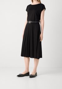 Mustat laskostetut hameet, jotka on yhdistetty tiukkaan mustaan paitaan, jota koristaa musta vyö. Jalkineet ovat mustat ballerinat, joissa on koristeellisia metalliosia.