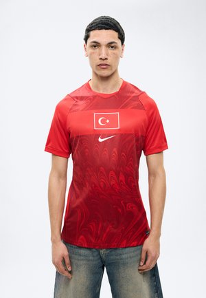 Ung mand iført en rød sportstrøje med det tyrkiske flag og Nike-logo, stående foran en ensfarvet hvid baggrund.