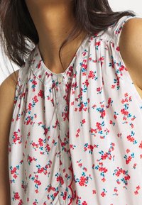 Blusa floral con una tela blanca adornada con patrones de flores rojas y azules, cuello redondo y detalles a botones en el frente.
